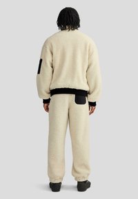 Completo giacca e pantaloni in pile color crema con polsini neri a coste, caratterizzato da una tasca sul retro dei pantaloni e una toppa nera sulla manica.