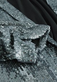 Silberner Paillettenstoff mit einer schimmernden Oberfläche, gegen einen schwarzen Textilhintergrund gefaltet. Verfügt über eine glatte Textur und unterschiedliche Lichtreflexionen.