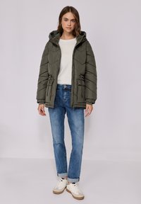 Doudoune vert olive avec capuche, cordons de tirage sur les côtés et texture matelassée. Associée à un jean bleu clair et des baskets blanches.