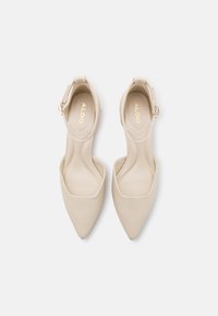 Escarpins beiges à talons hauts avec un bout pointu et une bride à la cheville, dotés d'un matériau lisse et d'un logo subtilement embossé sur la semelle intérieure.