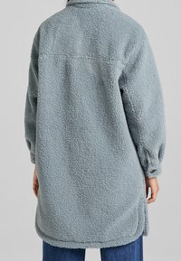 Light blue fleece jacket with long sleeves, textured fabric, and a collar. The design features a relaxed fit and side slits at the hem.