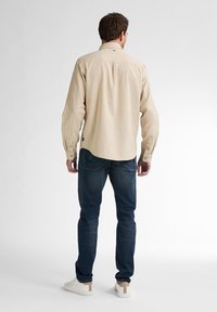 Lichtbeige langemou shirt met een rechte snit, knoopmanchetten, achterplooien en een klein logotag, gecombineerd met donkerblauwe jeans en witte sneakers.