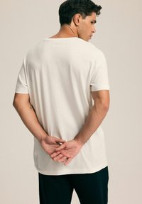 Biała bawełniana koszulka z luźnym krojem, krótkimi rękawami i okrągłym dekoltem, noszona przez osobę widzianą z tyłu, z rękami złożonymi.