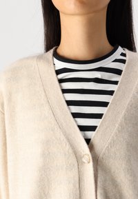 Beżowy sweter na drutach z dekoltem w serek i pojedynczym zapięciem na guzik, noszony na czarno-białej koszulce w paski. Miękka faktura i swobodny styl.