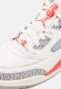 Gros plan sur une basket Air Jordan blanche et grise avec des accents rouges, des perles Paris Saint-Germain (PSG) sur les lacets et le logo Jumpman sur la languette et la perle.