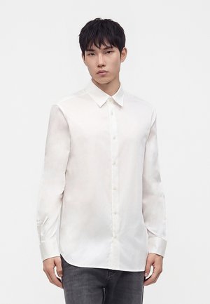 Jeune homme portant une chemise blanche unie à manches longues boutonnée et un jean gris foncé, debout devant un fond blanc.