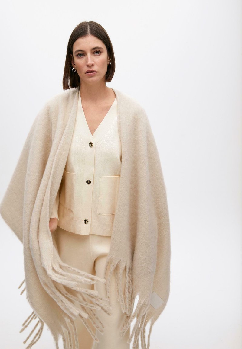 Bež šal s dugim resama, mekana tekstura, nosi se preko svijetle cardigan jakne s džepovima i crnim dugmadima. Jednostavan, minimalan dizajn.