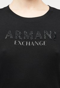 Melnā T-krekla ar teksturētu sudraba rakstu "ARMANI" un gludu metāla krāsas uzrakstu "EXCHANGE", klasiskā apaļā kakla dizainā.