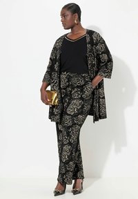 Černý květinový vzorovaný outfit se volnou kimono bundou, přiléhavými kalhotami a černým topem, doplněný zlatou kabelkou a černými podpatky.