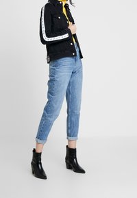 Chaqueta de mezclilla negra con acentos de rayas blancas, combinada con jeans azul claro de tirantes enrollados y botas de tobillo negras de punta afilada.