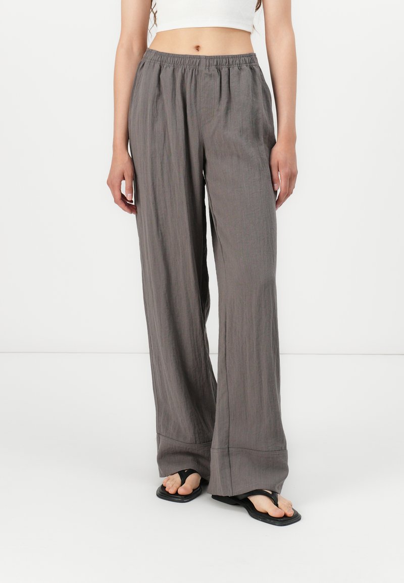 Pantalons larges gris avec une taille élastique, en tissu léger et avec une texture subtile. Associés à des sandales flip-flop noires.