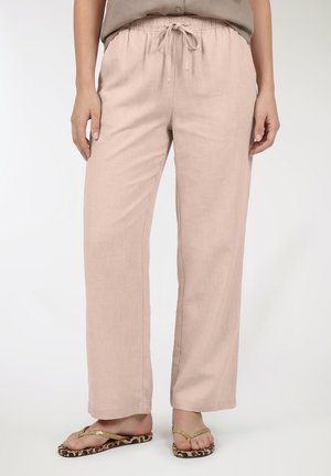 Donna con pantaloni beige con coulisse e infradito con stampa leopardata, in piedi con le mani rilassate ai lati contro uno sfondo semplice.