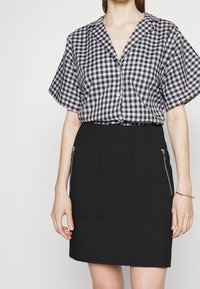 Gonna nera corta con tasche frontali e dettagli in zip argentati, abbinata a una camicia a maniche corte a quadri bianca e nera con bottoni.