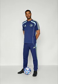 Mann i blå og lysegrønn Adidas sportsklær står med fotball nær føttene mot en ensfarget bakgrunn.