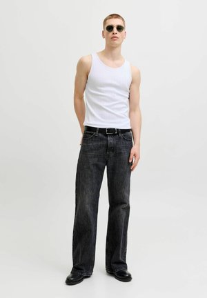 Fiatal férfi kerek napszemüvegben, fehér trikóban, fekete övvel, bő fekete farmerben és fekete cipőben, egyszerű világos háttér előtt áll.