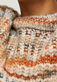 Gros plan sur le col d'un pull en tricot épais aux tons crème, orange, bleu et marron sur le cou et l'épaule d'une personne.