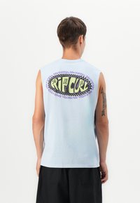 Man som bär en ärmlös ljusblå skjorta med en färgglad "Rip Curl"-logotyp och texten "Raw Energy" på ryggen, svarta shorts och en piltatuering på armen.