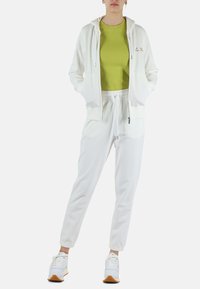 Felpa bianca con zip e tasche laterali, abbinata a una maglietta verde e pantaloni jogger bianchi. Le scarpe sono bianche con accenti multicolori e suole marroni.