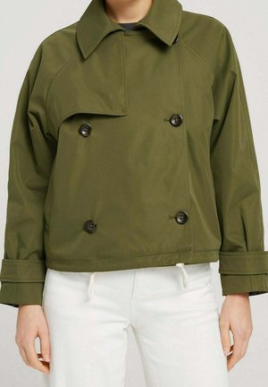 Personne portant une veste croisée vert olive avec un grand col et des boutons noirs, associée à un pantalon blanc.
