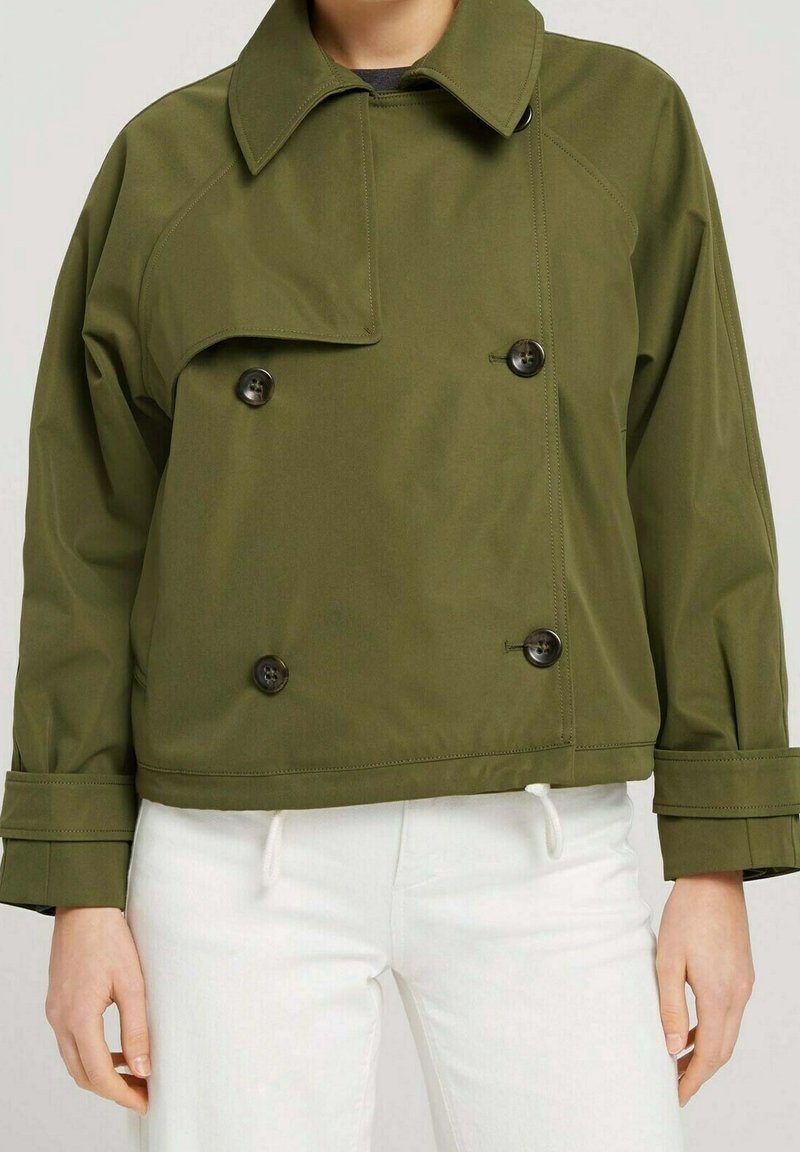 Personne portant une veste croisée vert olive avec un grand col et des boutons noirs, associée à un pantalon blanc.