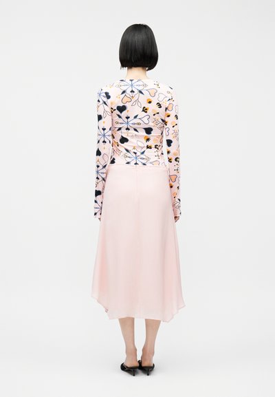 Femme aux cheveux courts noirs vue de dos, portant un haut à manches longues à motifs cœurs et fleurs, une jupe midi rose pâle et des talons noirs.