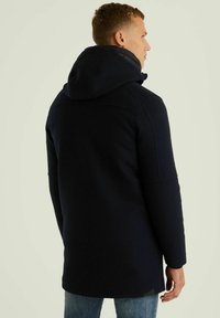 Tamsiai mėlynas su kapuce paltas su tekstūriniu audiniu, matomomis siūlėmis ir nugaroje esančia skeltimi. Turi laisvą siluetą ir derinamas su šviesiomis džinsinėmis kelnėmis.