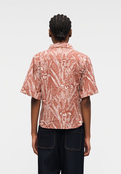 Personne aux cheveux tressés en chignon, portant une chemise à manches courtes de couleur rouille avec un imprimé botanique blanc et un pantalon foncé, vue de dos.