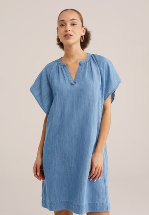 Nő világoskék, bő szabású farmerruhában, rövid, széles ujjakkal és V-nyakkivágással, egyszínű bézs háttér előtt áll.