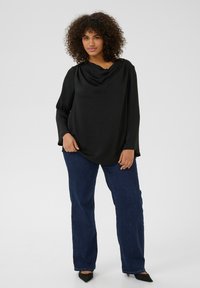 Sort, draperet bluse lavet af glat stof, med lange ærmer; parret med mørkeblå, lige ben jeans og spidse sorte sko.