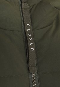 Chaqueta acolchada de color verde oscuro con una cremallera central que tiene una lengüeta texturizada con la etiqueta "CLOSED" en blanco, material liso de poliéster y diseño acolchado.