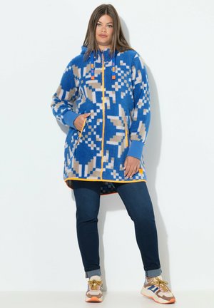 Fleece takki sinisellä, vaaleanbeigeällä ja oranssilla geometrisella kuviolla. Varustettu vetskarilla, hupulla ja taskuilla. Yhdistetty tummiin farkkuihin ja lenkkareihin.