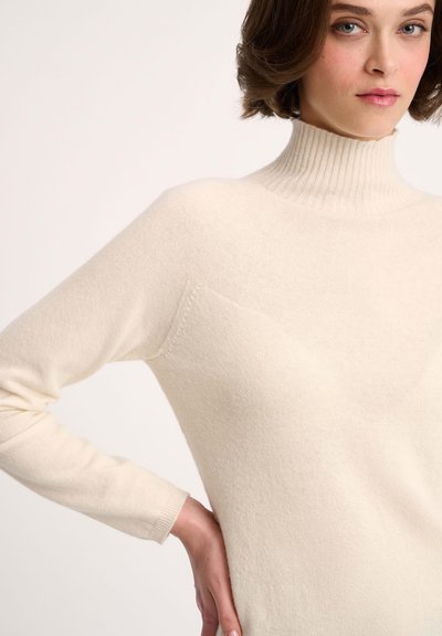 Pulover turtleneck de culoare bej deschis, realizat dintr-o țesătură moale, tricotată. Are mâneci lungi și un buzunar discret pe partea stângă.