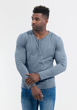 Langärmliges Henley-Shirt aus weichem blauen Stoff mit rundem Ausschnitt, Frontknöpfen und taillierten Ärmeln. Kombiniert mit blauen Denim-Jeans.