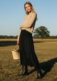 Beige pulóver, fekete, rétegelt maxi szoknya, magas fekete bőr csizma és fonott kosár táska. A modell egy mezőn áll, tiszta kék égbolt alatt.