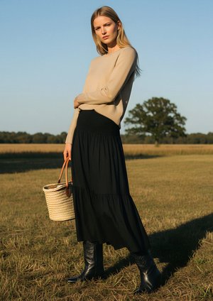 Beige trui, zwarte gelaagde maxi-rok, hoge zwarte leren laarzen en een geweven mandtas. Model staat in een veld onder een heldere blauwe lucht.