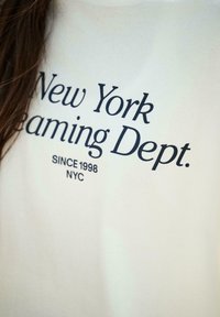 Fehér pamut póló, amelyen nagy fekete betűkkel szerepel a "New York Dreaming Dept." felirat, alatta pedig a "Since 1998 NYC" szöveg.