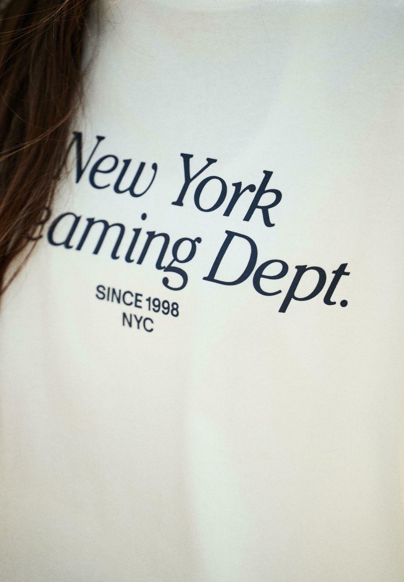 Fehér pamut póló, amelyen nagy fekete betűkkel szerepel a "New York Dreaming Dept." felirat, alatta pedig a "Since 1998 NYC" szöveg.