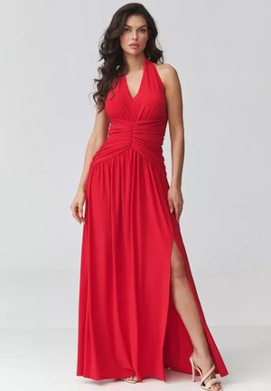 Kobieta ubrana w czerwoną suknię bez rękawów z dekoltem halter, marszczoną talią i rozcięciem do uda, połączoną z beżowymi sandałami na wysokim obcasie, stojąca.