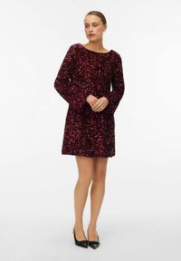 Korte, bordeauxrode jurk met lange, flared mouwen, versierd met pailletten voor textuur en glans. Gestyled met puntige zwarte hakken.