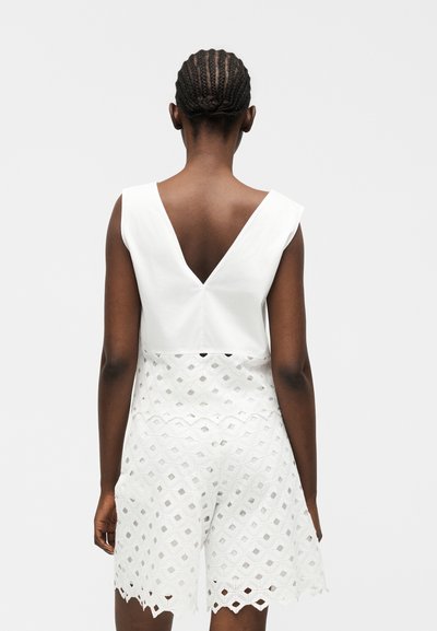 Γυναίκα με κοτσίδες, που φοράει ένα λευκό αμάνικο φόρεμα με βαθύ V στο πίσω μέρος και γεωμετρικό δαντελωτό σχέδιο στη φούστα.