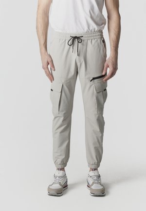Hombre vistiendo pantalones cargo gris claro con puños elásticos, cintura ajustable con cordón negro, bolsillo con cremallera y zapatillas blancas con detalles en beige y rojo.