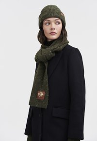 Mørkegrøn strikket halstørklæde med plettede accenter og et lædermærke. Brugt sammen med en matchende strikket hue og en sort frakke.