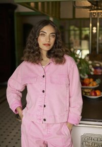 Roza jeans jakna z dvema žepoma na prsih, črnimi gumbi in usklajenimi hlačami. Teksturirana tkanina in sproščen kroj, prikazano v svetlem notranjem okolju.