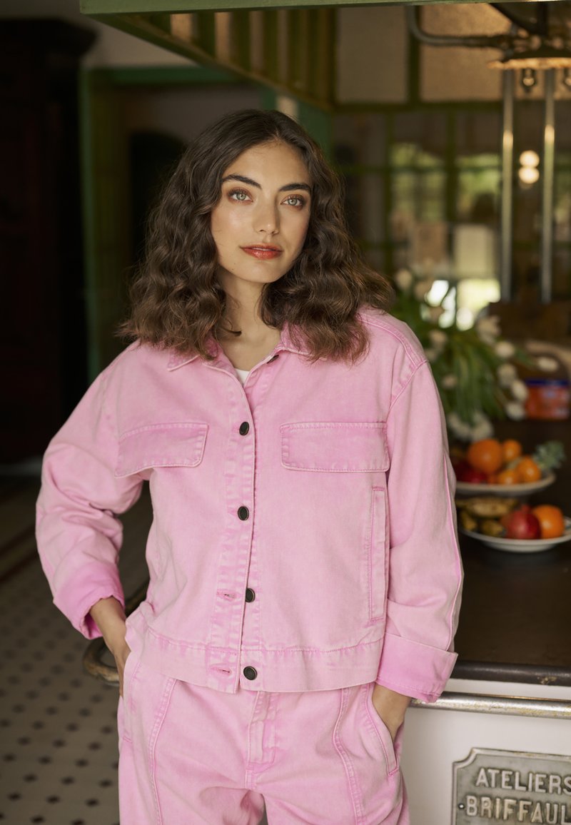 Roza jeans jakna z dvema žepoma na prsih, črnimi gumbi in usklajenimi hlačami. Teksturirana tkanina in sproščen kroj, prikazano v svetlem notranjem okolju.