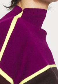 Gebreide trui met een hogere hals in paars met gele en bruine accenten. De textuur is glad, met contrasterende kleurstroken.