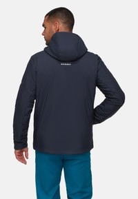 Uomo in giacca a cappuccio blu navy con logo "Mammut" sulla schiena e pantaloni turchesi, di spalle con una mano sui fianchi.