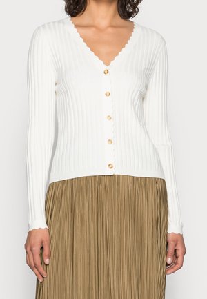 Femme portant un cardigan blanc côtelé boutonné avec des bords festonnés et une jupe plissée marron clair.