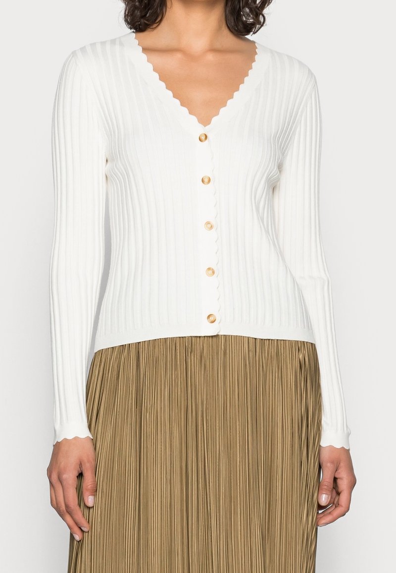 Femme portant un cardigan blanc côtelé boutonné avec des bords festonnés et une jupe plissée marron clair.