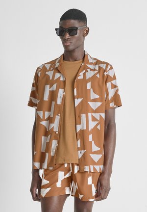 Man draagt een bruin-wit geometrisch patroon kortärmelig overhemd en bijpassende korte broek met een bruin T-shirt en zwarte zonnebril.