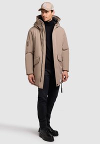 Parka acolchada beige con capucha, cierre frontal de cremallera y botones a presión, dos bolsillos delanteros, usada sobre un cuello de tortuga negro y jeans negros, completada con botas negras.